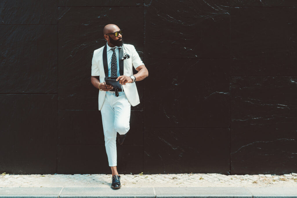 A stylish bearded man in a white fashion business suit and sunglasses is leaning against a black marble wall, holding his clutch and looking aside, with a copy space place on the right
