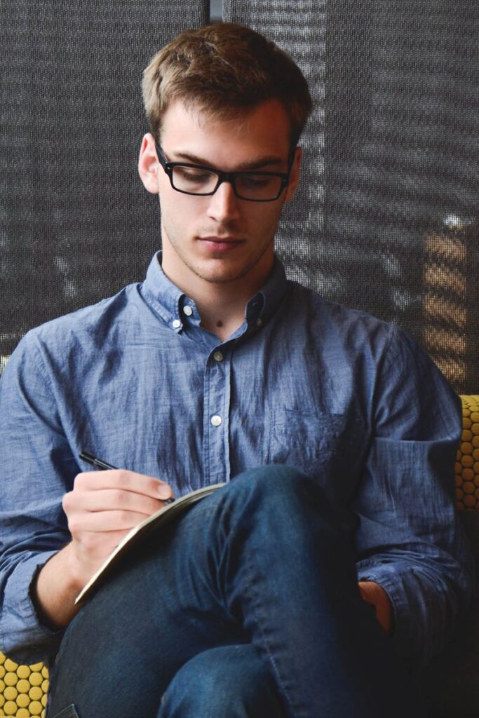 entrepreneur-startup-start-up-man-39866-39866 A young man in glasses writes in a notebook while sitting on a stylish couch indoors.