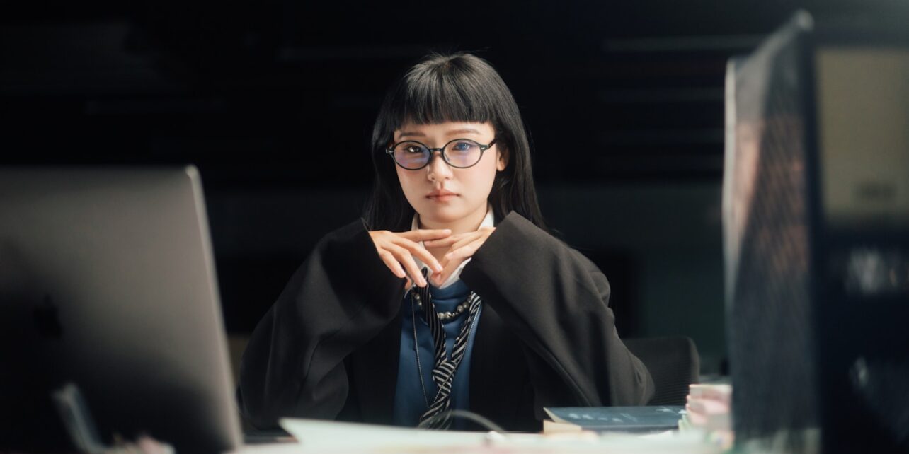 Woman looks intently at the camera, working on computers.